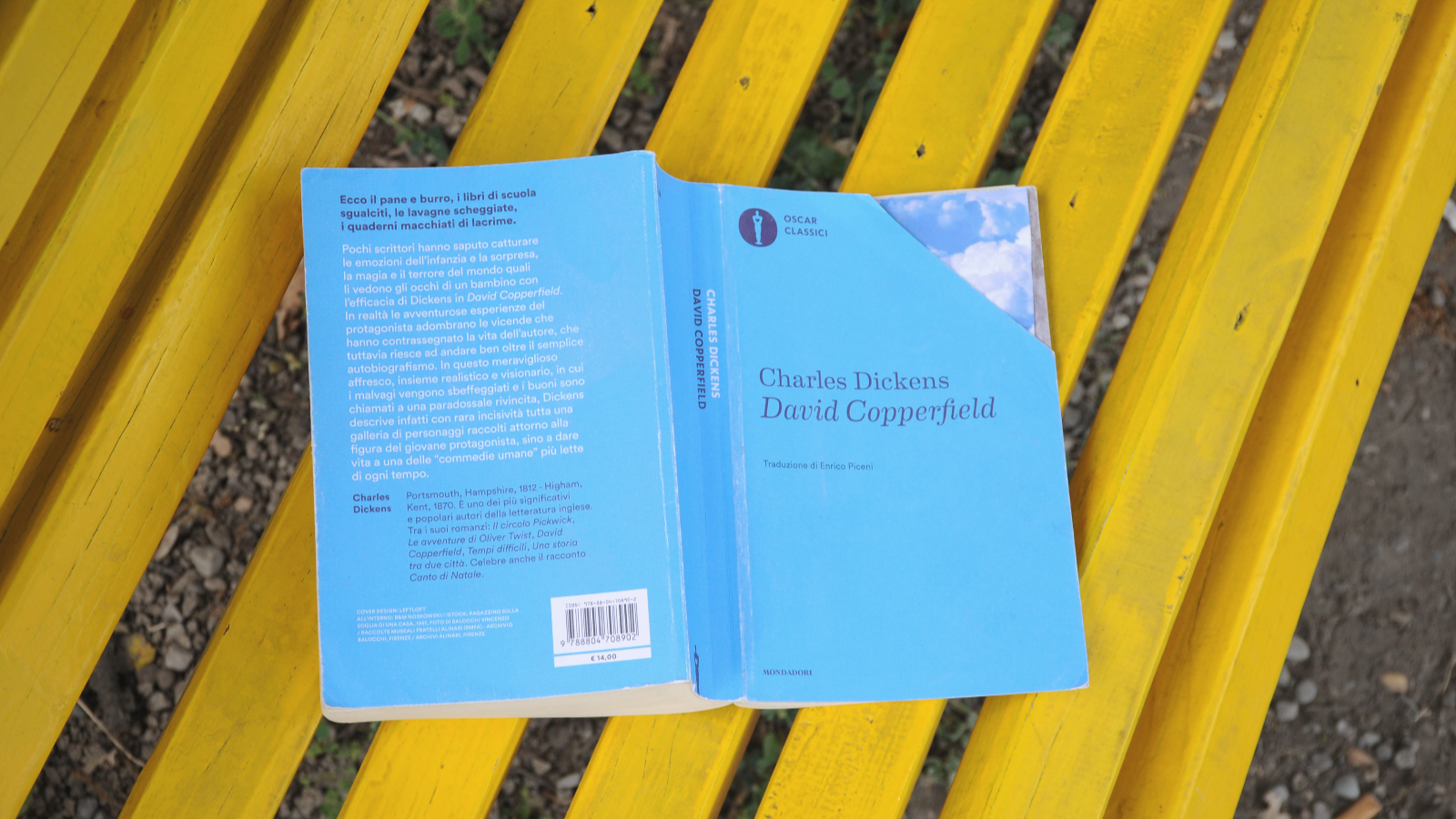 il libro, poggiato sulle pagine aperte, si trova su una panchina gialla, sull'immagine si trova il titolo del libro, ovvero David Copperfield, edito nel 1850 e scritto da Charles Dickens